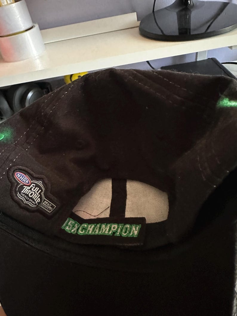 May include: A dark brown baseball cap featuring an embroidered NHRA Full Throttle Energy Drink logo on the side and a green embroidered patch on the back strap that reads 15x CHAMPION in capital letters with a white interior lining.
