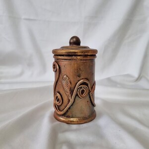 May include: A small, round, copper-coloured jar with a lid. The jar is decorated with a vine-like design with leaves.