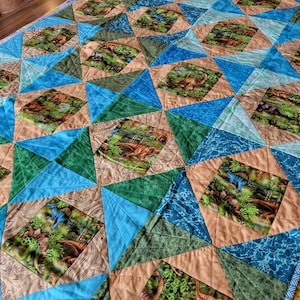 May include: A colorful quilt featuring a repeating pattern of dinosaur images set within diamond and triangle shapes. The quilt uses shades of blue, green, and tan. The dinosaur images are set in a jungle scene.