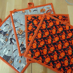May include: Three square quilted pot holders featuring Halloween themes. One displays a gray background with mummies, pumpkins, and haunted houses. The other two feature a repeating orange and black grid pattern with pumpkins and black cats.