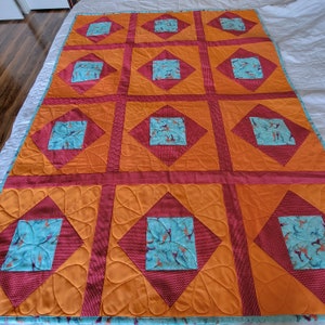 Könnte beinhalten: Handgefertigte Steppdecke mit lebendigem Design. Die Steppdecke hat ein Rastermuster aus orangefarbenen Quadraten mit roten Diamantakzenten. Jeder Diamant enthält ein türkisfarbenes Quadrat mit einem Muster. Ein gemütliches Wohnaccessoire.