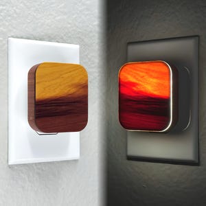 May include: A wooden nightlight with a sunset design. The nightlight is plugged into a standard electrical outlet. The nightlight is lit up, showing the sunset design in a warm orange and red color.