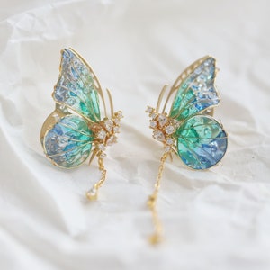 May include: A pair of gold butterfly earrings with blue and green iridescent wings and clear crystal accents. The earrings have a dangling chain with a single crystal at the end.