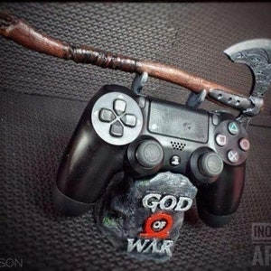 Puede incluir: Un mando de PlayStation 4 negro con un soporte personalizado que presenta el logotipo de God of War. El soporte está diseñado para parecerse al Hacha Leviathan del juego, con un mango de madera y una cabeza de hacha plateada.