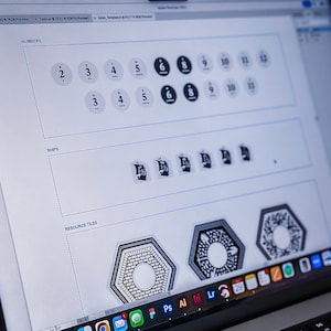 May include: A computer screen displaying a design program with a white background. The screen shows a design for a board game with numbers, ships, and resource tiles. The text "NUMBERS", "SHIPS", and "RESOURCE TILES" is visible on the screen.