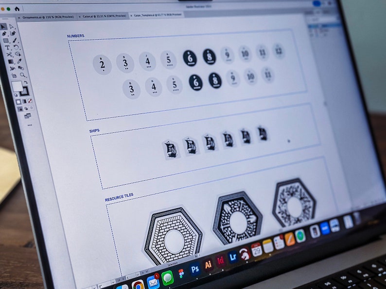May include: A computer screen displaying a design program with a white background. The screen shows a design for a board game with numbers, ships, and resource tiles. The text "NUMBERS", "SHIPS", and "RESOURCE TILES" is visible on the screen.