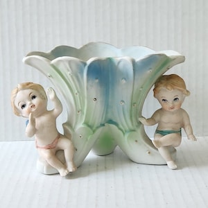 Peut inclure: Un pot en céramique blanche en forme de fleur avec deux figurines de chérubins de chaque côté. Le pot est décoré d'accents bleus, verts et dorés.