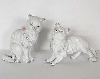 May include: Two white porcelain cat figurines with pink flower accents. The cat on the left is sitting with its tail curled around its legs. The cat on the right is lying down with its head turned to the side.