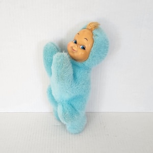 May include: A blue plush toy with a smiling face and blonde hair. The toy is wearing a blue jumpsuit and has large blue eyes.