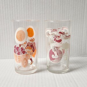May include: Two clear glass tumblers with colourful illustrations. One tumbler features a person pushing a cart full of red and orange fruit. The other tumbler features a person riding a bicycle with a red and white design.
