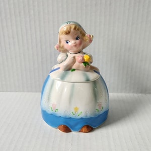 Peut inclure: Un sucrier en céramique vintage en forme de fille portant une robe bleue et blanche avec une fleur jaune. La fille a les cheveux blonds et les yeux bleus.