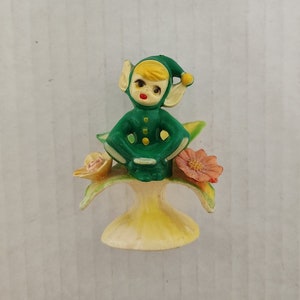 Peut inclure: Une figurine d'elfe verte avec un chapeau jaune et une bouche rouge est assise sur une fleur jaune avec des pétales roses. L'elfe tient une tasse verte. La figurine est sur une base jaune.