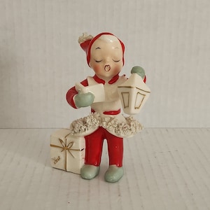 Peut inclure: Une figurine en porcelaine vintage d'un lutin de Noël tenant une lanterne et une boîte à cadeaux. Le lutin porte un costume rouge et blanc avec une bordure en fourrure blanche. La lanterne est dorée avec une bougie blanche à l'intérieur. La boîte à cadeaux est blanche avec un ruban doré et les mots "Joyeux Noël" écrits dessus.