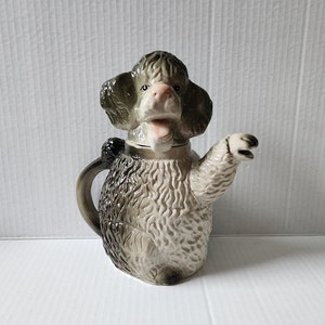 May include: A ceramic teapot shaped like a poodle with a brown and white fur pattern. The teapot has a spout shaped like a paw and a lid shaped like the poodle's head.