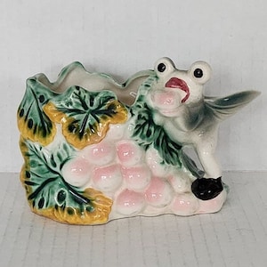 May include: A ceramic planter shaped like a frog with a green, yellow and pink grapevine design. The frog has a wide open mouth and large eyes.