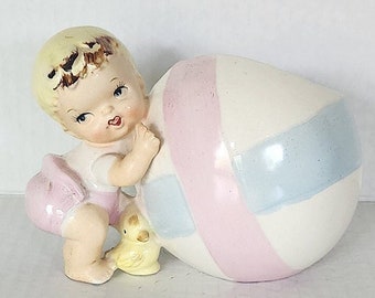May include: A vintage ceramic figurine of a baby in a pink outfit, pushing a large, egg-shaped object. The egg is white with pink and blue stripes. The baby has blonde hair and a sweet expression.