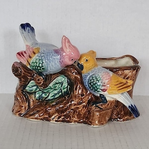 May include: A ceramic planter shaped like a tree trunk with two birds perched on it. The birds are painted in shades of blue, pink, yellow, and green. The planter is a great way to add a touch of nature to your home.