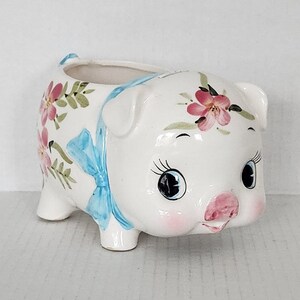 May include: A white ceramic piggy bank planter with blue and pink floral designs. The piggy bank has a large snout and big eyes.