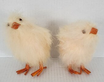 May include: Two fluffy white chicks with orange beaks and legs. The chicks are facing different directions and are standing on a white surface.