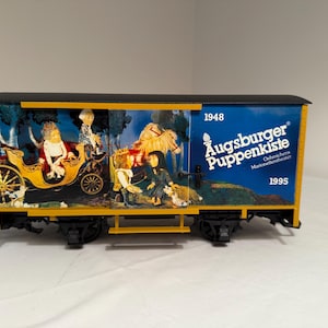 May include: A model train car with a black roof and yellow frame. The sides feature a colorful illustration of a horse-drawn carriage and figures. The text "Augsburger Puppenkiste" is displayed on a blue background, with the years 1948 and 1995.