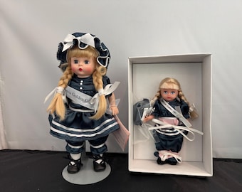 May include: Two porcelain dolls dressed in blue and white sailor outfits. The larger doll is standing on a white stand and has blonde braids. The smaller doll is in a box and has blonde pigtails. Both dolls have a white sash with the text "Miss Golden Girl".