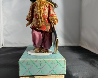 May include: A vintage Madame Alexander doll dressed in a traditional Turkish costume. The doll is wearing a red fez hat, a gold and red patterned jacket, purple trousers, and gold shoes. The doll is holding a book and is standing on a blue and white box. The box has the words "Madame Alexander" printed on it, along with the words "Turkey 787".