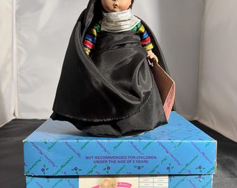 May include: A black and colorful doll wearing a black dress and a silver necklace. The doll has brown hair and brown eyes. The doll is sitting on a blue box with the text "Madame Alexander" and "NOT RECOMMENDED FOR CHILDREN UNDER THE AGE OF 3 YEARS".