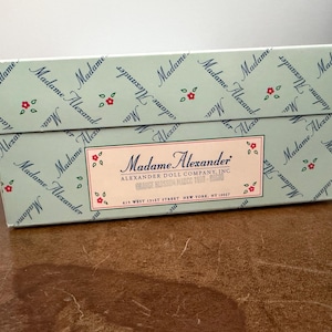 May include: A rectangular box with a light blue background and the words "Madame Alexander" in a repeating pattern. The box has a label with the words "Madame Alexander" and "Alexander Doll Company, Inc."