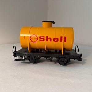 May include: A yellow Shell oil tank car model with the Shell logo in red. The tank car sits on a black flatbed with black wheels. The model is a miniature replica of a real-life train car.