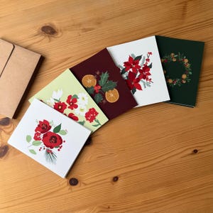 May include: A collection of Christmas greeting cards with floral and botanical designs. The cards feature illustrations of poinsettias, holly, and orange slices. The cards are arranged on a wooden surface, with a brown envelope visible.