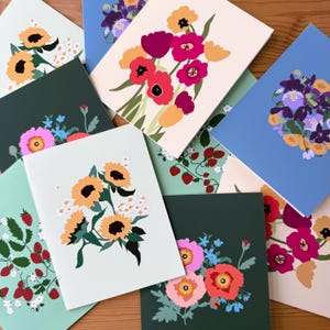 May include: A set of eight blank greeting cards featuring colorful floral illustrations. The cards are printed on white cardstock and have a matte finish. The illustrations include sunflowers, poppies, and other wildflowers.