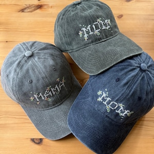 May include: Three baseball caps in shades of grey and blue are displayed. Each cap has the word "MOM" embroidered in a colourful, cross-stitch style. The caps are arranged on a light brown wooden surface.