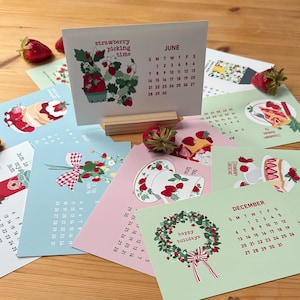 May include: A desk calendar with monthly pages featuring strawberry-themed illustrations. The June page is displayed, showing a strawberry plant graphic and a calendar grid. Other pages show strawberry desserts and floral arrangements.