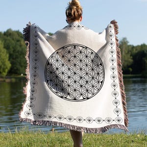 May include: A cream-colored throw blanket with a large black geometric design in the center. The blanket has a fringe border and a repeating circular pattern along the edges. The person is standing near a lake.