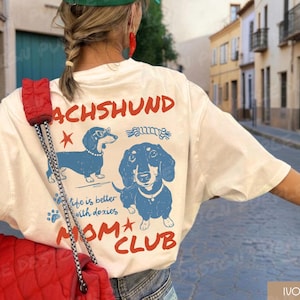 May include: Ivory-colored t-shirt with a dachshund graphic in blue and the text "DACHSHUND MOM CLUB." The shirt features two dachshund illustrations, one wearing glasses. The phrase "life is better with doxies" is also printed on the shirt.