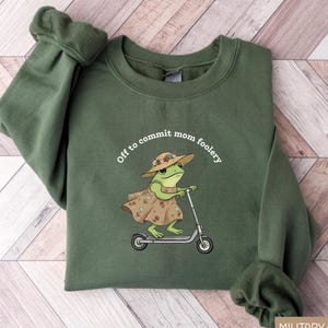 May include: Military green sweatshirt with the phrase "Off to commit mom foolery" and a graphic of a frog riding a scooter. The frog wears a floral dress and hat. The sweatshirt has long sleeves.