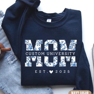 May include: Navy blue sweatshirt with the word "MOM" in a floral design, along with "CUSTOM UNIVERSITY MUM" and "EST. 2025". A coffee mug, jeans, and a sneaker are also in the image.