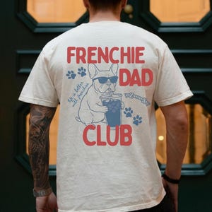 May include: Ivory t-shirt with a graphic of a French bulldog wearing sunglasses, holding a drink, and the words "FRENCHIE DAD CLUB" in red. Blue paw prints and the phrase "life is better with frenchies" are also included on the shirt.