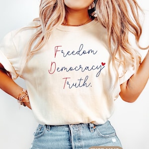 May include: Ivory-colored t-shirt with the words "Freedom, Democracy, Truth" in red and blue script. A small red heart is next to the word "Democracy". The shirt is paired with blue jeans. The text "IVORY COLOR" is at the bottom.