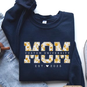 May include: Navy blue sweatshirt with the text "MOM CUSTOM UNIVERSITY" in a floral design. The sweatshirt also includes "EST. 2025" and a heart symbol. A coffee mug, jeans, and a sneaker are in the background.