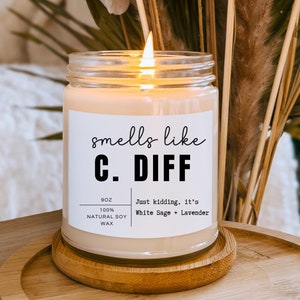 May include: A lit, clear glass jar candle on a wooden coaster. The candle has a white label with the text "smells like C. DIFF" and "Just kidding, it's White Sage + Lavender." The candle is 9 oz and made of natural soy wax.