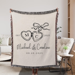 May include: Cream-colored throw blanket with black fringe and text. The blanket features two connected heart shapes with the letters "M" and "C", a bow, the names "Michael & Caroline", the date "08.28.2025", and the word "EST. 2025".