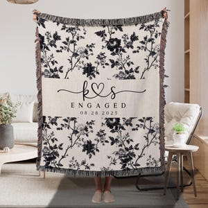 May include: A cream-colored throw blanket with a black floral pattern and fringe. The blanket features the text "Engaged" and the date "08.28.2025" with a heart symbol. The initials "k&s" are also displayed.