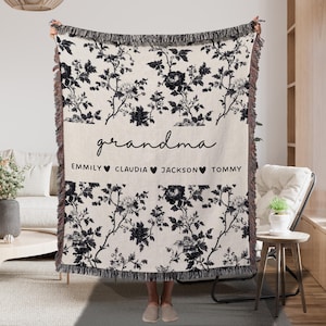 May include: A cream-colored throw blanket with a black floral pattern and fringe. The word "grandma" is written in script, with names below. A cozy home decor item.