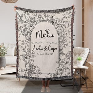 May include: A cream-colored throw blanket with a floral design and fringe trim. The blanket features a central arch with the names "Miller" and "Amelia & Cooper" and the date "August 18, 2025". The floral pattern includes various flowers and leaves.