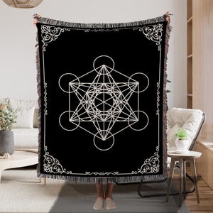 May include: Black throw blanket with a white geometric Metatron's Cube design. The blanket has a decorative border and fringe trim. The design features interconnected circles and lines, creating a symmetrical pattern.