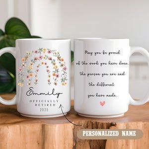 Peut inclure: Une tasse &agrave; caf&eacute; en c&eacute;ramique blanche avec un motif floral et le texte "Emmily Officially Retired 2025". L'autre c&ocirc;t&eacute; de la tasse a le texte "May you be proud of the work you have done. The person you are and the different you have made." avec un petit c&oelig;ur rose.
