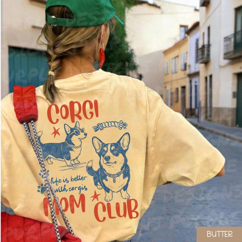 Puede incluir: Camiseta de color mantequilla con un gr&aacute;fico de dos perros corgi y las palabras "CORGI" y "MOM CLUB" en rojo. El texto "life is better with corgis" tambi&eacute;n est&aacute; impreso en la camiseta. La camiseta la lleva una persona.