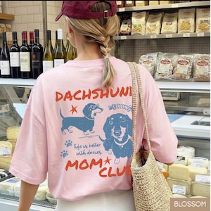 May include: A light pink t-shirt with a dachshund graphic and the words "DACHSHUNI MOM CLUB". The graphic features two blue dachshunds and the phrase "life is better with doxies".
