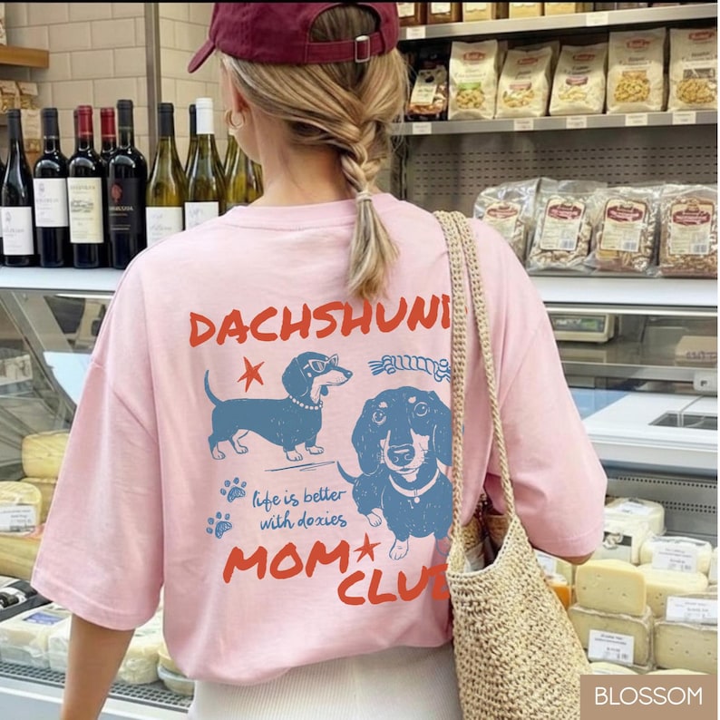 May include: A light pink t-shirt with a dachshund graphic and the words "DACHSHUNI MOM CLUB". The graphic features two blue dachshunds and the phrase "life is better with doxies".